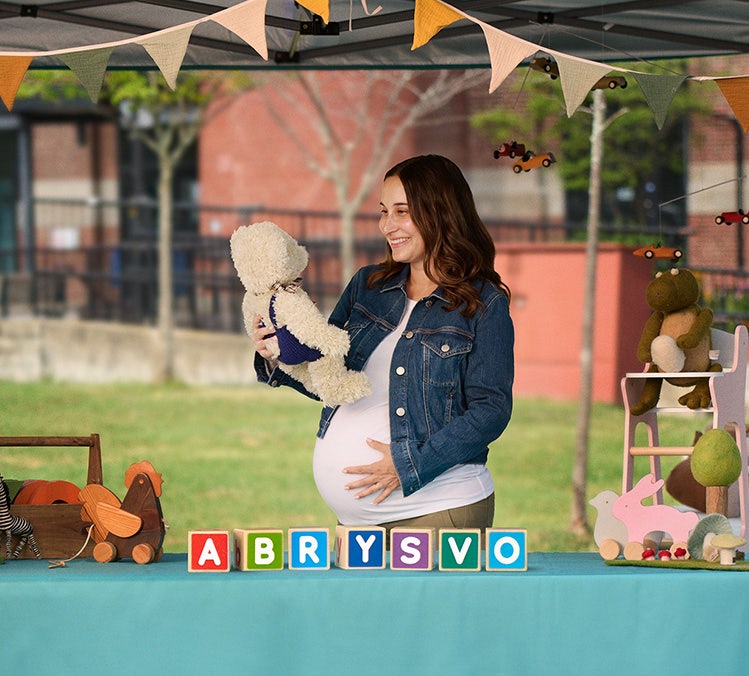 Personne enceinte entourée d’articles pour bébé et d’un présentoir ABRYSVOMD, illustrant la vaccination maternelle contre le VRS pendant la grossesse.